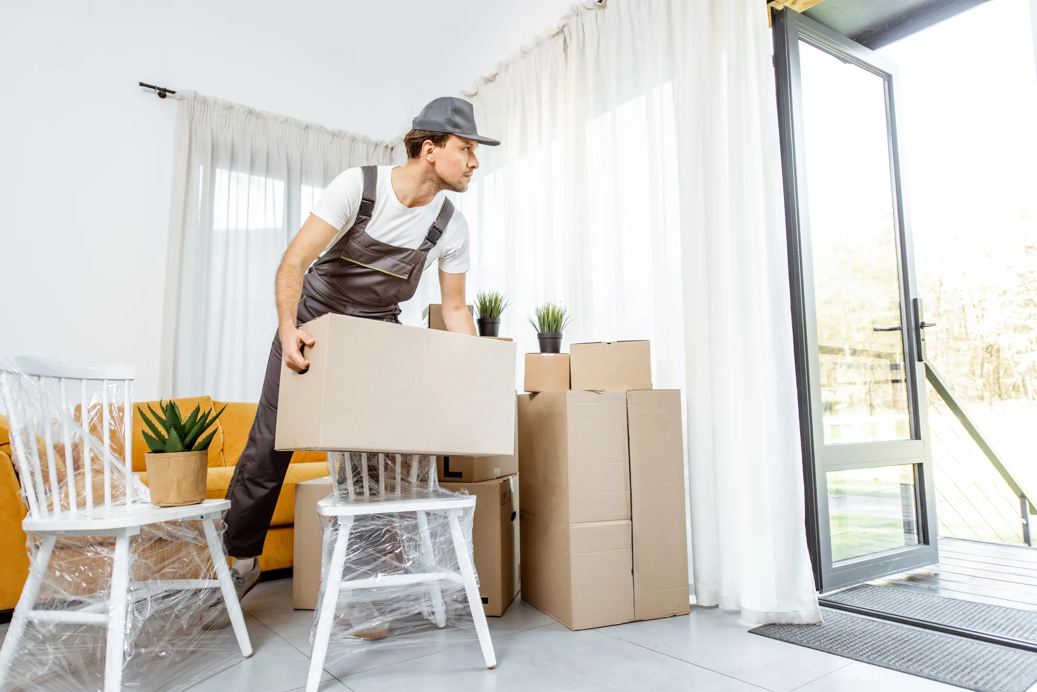Handsome mover in workwear performing professional delivery of a goods and furniture during relocation process to a new house, interior view