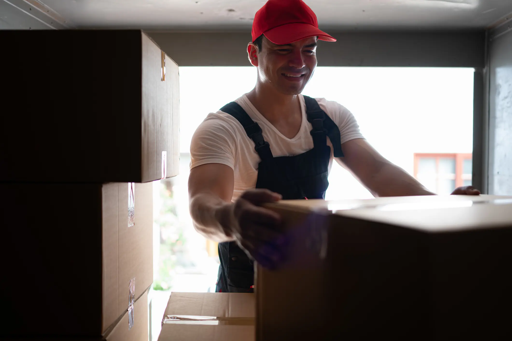 Handsome young delivery man in red cap and gloves holding a box in the truck