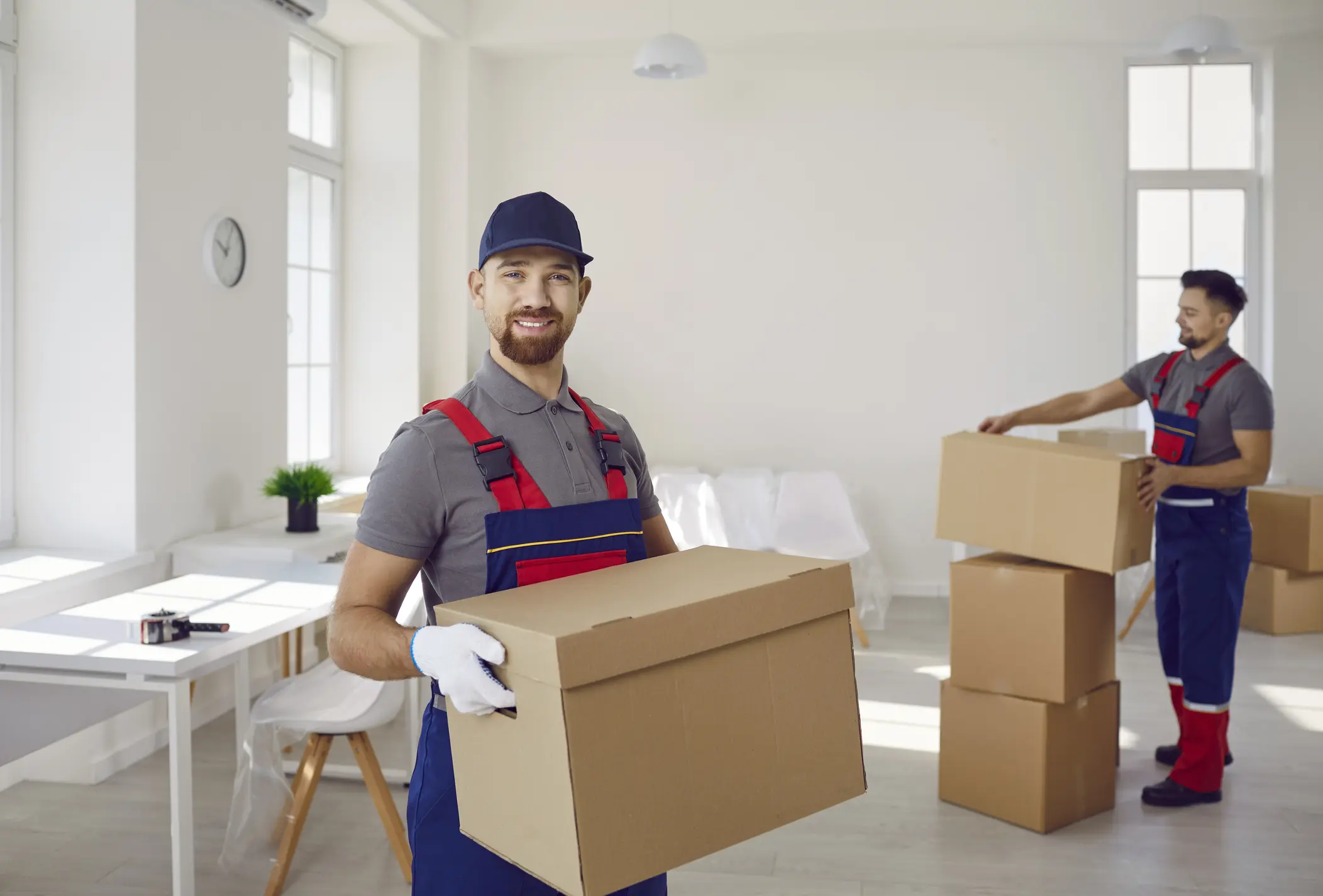 Portrait of smiling young man mover worker in uniform holding a cardboard box and looking cheerful at camera standing in empty new apartment. Move, relocation and moving service concept.