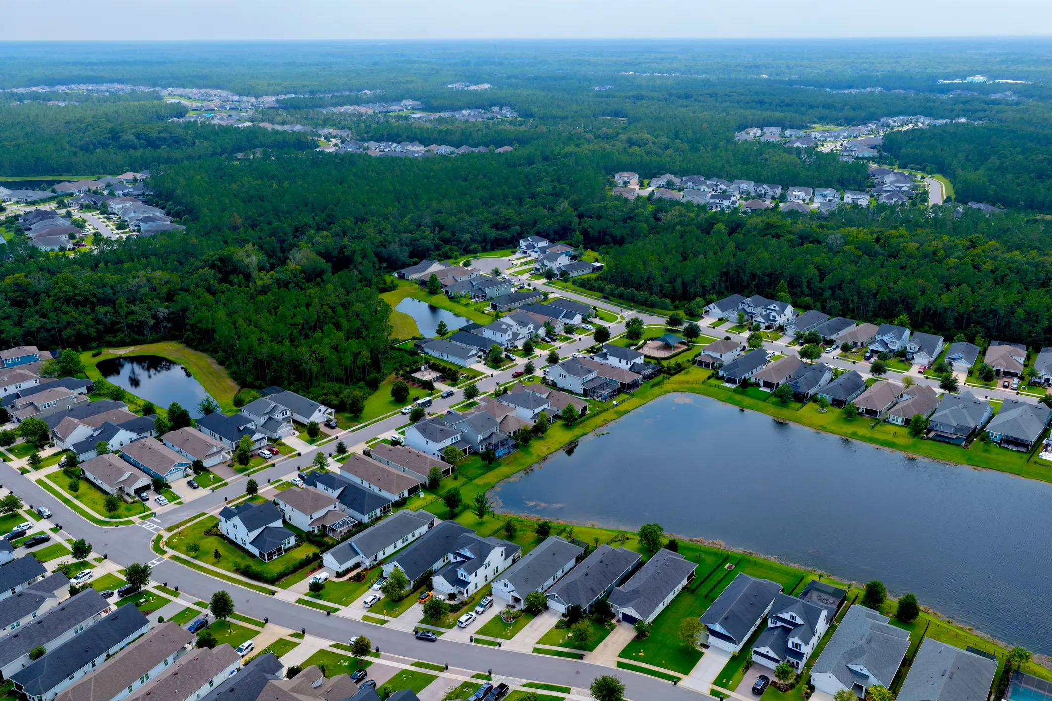 This aerial photo features a large subdivision of single-family homes in Nocatee, Florida. Retention ponds are also visible. Nocatee is a massive, award-winning master-planned coastal community in Ponte Vedra. Home prices typically range from the $400s to over $2 million, and residents enjoy a vibrant golf-cart lifestyle with access to water parks, nature preserves, and top-rated schools. Many of the homes include open-concept living, energy-efficient designs (100% Energy Star), pools, and lanais. Retention ponds are used to control water flow and flooding in the development's residential neighborhoods.