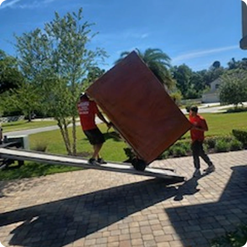 Two men in red shirts unloading a large piece of furniture from a moving truck