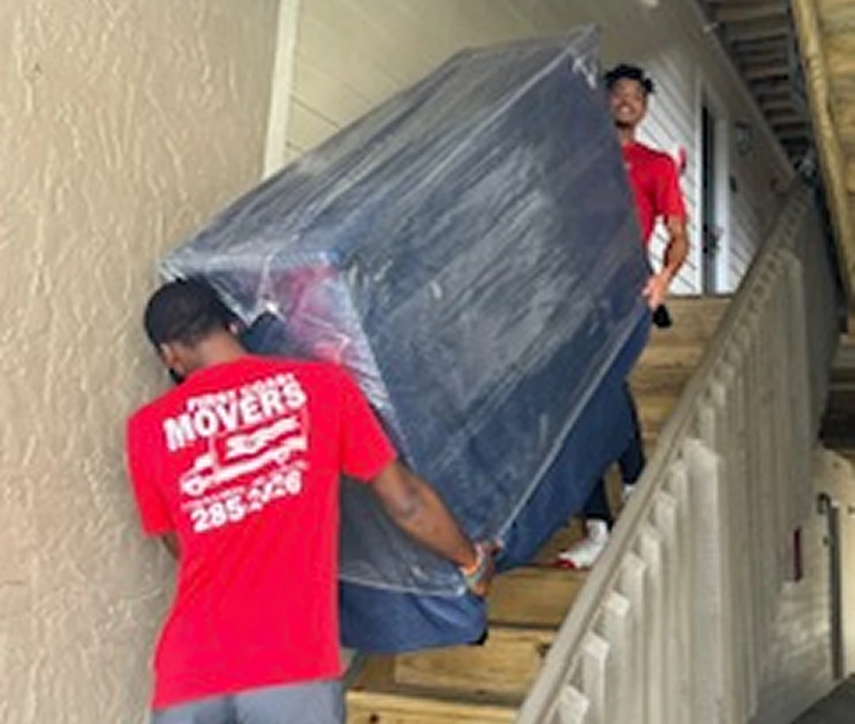 Two movers in red shirts carrying a large piece of furniture up the stairs of a building